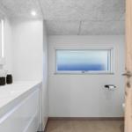 Badezimmer mit Waschtisch, Spiegel und Fenster. Holztür und Toilettenpapierhalter sichtbar.
