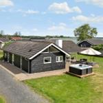 Black wooden house with hot tub and terrace. Lawn and water in the background.