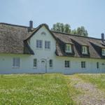 Weißes Haus mit strohgedecktem Dach und mehreren Fenstern in grüner Umgebung.