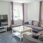 Wohnzimmer mit Sofa, Glas-Tisch und Fernseher. Fenster mit Vorhängen.
