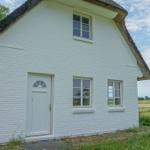 Weißes Haus mit Strohdach, Fenstern und Tür. Grün umgeben, Felder im Hintergrund.