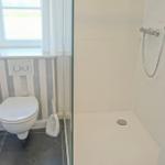 Badezimmer mit Dusche, Toilette und Fenster mit Vorhängen.