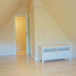 Zimmer mit schrägen Wänden, weißem Schrank und Holzboden. Tür führt in einen anderen Raum.