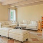 Wohnzimmer mit weißen Sofas, Holzbalken und Terrassentür. Kamin und TV-Tisch sichtbar.