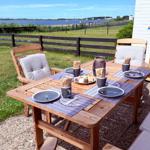 Holztisch mit Stühlen und Tischdecken auf Terrasse mit Blick auf Wasser und Windräder.