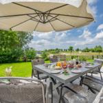 Terrasse mit Tisch, Stühlen und Sonnenschirm. Frühstückszubehör und Blick auf Grünflächen.