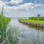 Wasserkanal mit Gräsern und hölzernem Ufer, Windfahne und Straßenschild