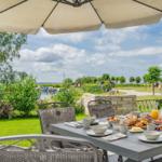 Terrasse mit Tisch, Stühlen und Sonnenschirm. Frühstück auf dem Tisch. Blick auf Wasser und Grün.