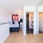 Schlafzimmer mit Bett, Wandbild und Tür zum Bad. Holzboden und Spiegeltür sichtbar.