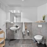 Badezimmer mit Waschbecken, Toilette und Holzregal. Graue Fliesen und Spiegel.