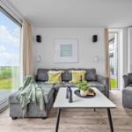 Wohnzimmer mit grauem Sofa, Tisch und Blick auf Wasser.