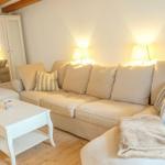 Wohnzimmer mit Ecksofa, Beistelltisch und zwei Stehlampen. Holzboden und weißer Kleiderschrank.