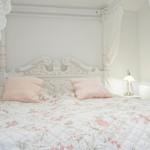 Schlafzimmer mit weißem Bett, floraler Bettwäsche und weißen Vorhängen.
