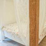 Schlafzimmer mit Bett unter Baldachin, Nachttisch und Fenster mit floraler Gardine.