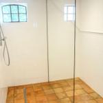 Duschkabine mit Glaswand, weißen Wänden und Terrakottbodenfliesen. Kleines Fenster mit mehreren Glasfeldern.