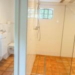 Badezimmer mit Dusche, Toilette und Waschbecken. Terrakottafußboden und Fenster mit Gitter.