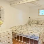 Schlafzimmer mit weißem Bett, Schrank und Nachttisch. Holzbalken und Fenster sichtbar.
