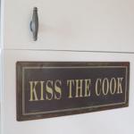 Schild mit der Aufschrift KISS THE COOK an einer Küchenwand.