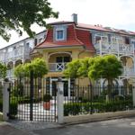 Haus mit rotem Dach und weißen Balkonen hinter schwarzem Zaun.