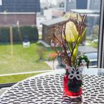 A tulip in a red glass vase sits on a table by a window overlooking a garden.