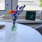 Auf dem Tisch steht eine Vase mit Blumen. Im Hintergrund sind Fenster und ein Sessel zu sehen.