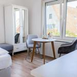 Schlafzimmer mit Bett, Kleiderschrank und Tisch. Fenster zeigt Dach und Grün.