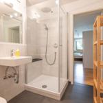 Badezimmer mit Dusche, Waschbecken und Holzregal.