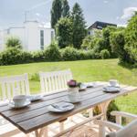 Holztisch mit weißen Stühlen auf Terrasse, Blick auf grünen Rasen und Bäume.