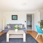 Wohnzimmer mit Sofa, Tisch und Stuhl. Durchgang zur Küche. Wandbild und Pflanze.