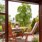 Terrasse mit Holztisch, Stühlen und Blumenarrangement. Blick auf grüne Bäume und Garten.
