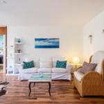 Wohnzimmer mit Sofa, Sessel und Holzboden. Küche im Hintergrund sichtbar.