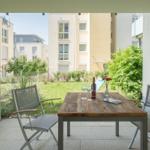 Terrasse mit Tisch und zwei Stühlen. Hintergrund: Grünfläche und Wohnhäuser.