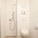 Dusche mit Duschbrause, WC und Toilettenpapierhalter in weißer Fliesenwand.