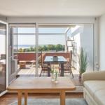 Wohnraum mit Balkonblick auf Wasser und Grün. Holztisch, Sofa, Glastüren.