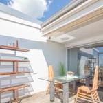 Terrasse mit Glas-Tisch, Holzstühlen und Metallregal an weißer Wand. Durchgehende Glastür zum Innenraum.