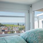 Zimmer mit Blick auf grüne Hügel und Häuser. Durchgehendes Fenster und Balkontür.