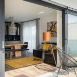 Wohnbereich mit Sofa, Stehlampe und Terrasse mit Stühlen. Durchgehende Glasfront.