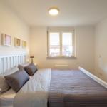 Schlafzimmer mit weißem Bett, Fenster und Nachttisch mit Lampe.