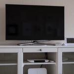 Flachbildfernseher auf weißem Sideboard mit Telefon und Digitaluhr.