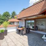 Terrasse mit Tisch, Stühlen und Pflanzen. Blick ins Haus mit Wohnzimmer und Glasfront.