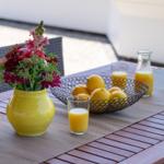 Außentisch mit Blumen, Orangensaft und Zitronen. Holzplatte und Stühle sichtbar.