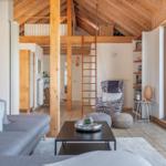 Wohnbereich mit Sofa, Couchtisch und Sessel unter Holzdecke. Treppenhaus und Küchenbereich sichtbar.
