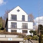 Weiße Villa mit Balkonen und Dachfenster, umgeben von Bäumen und Grünflächen.