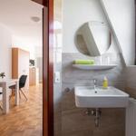 Badezimmer mit Waschbecken, Spiegel und Fenster. Im Hintergrund ist ein Essbereich mit Tisch und Stühlen.