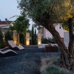 Garten mit Liegestühlen, Feuerkorb und Olivenbaum. Pool und Beleuchtung im Hintergrund.