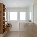 Badezimmer mit Dusche, Waschbecken und Heizung. Holzregal und Fenster sichtbar.