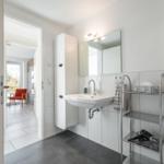 Badezimmer mit Waschbecken, Spiegel und Dusche. Durchgang zu Wohnbereich mit rotem Sessel.