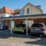Zwei Fahrzeuge unter einem Carport vor einem Haus mit rotem Dach.