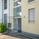 Eingangsbereich mit Glasfront und Briefkasten neben grüner Hecke.