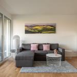 Wohnzimmer mit grauem Sofa, rosa Kissen und großem Landschaftsbild an der Wand.